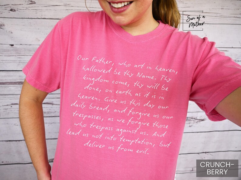 May include: Pink t-shirt with white text that reads "Our Father, who art in heaven, hallowed be thy Name. Thy kingdom come, thy will be done, on earth as it is in heaven. Give us this day our daily bread, and forgive us our trespasses, as we forgive those who trespass against us. And lead us not into temptation, but deliver us from evil."