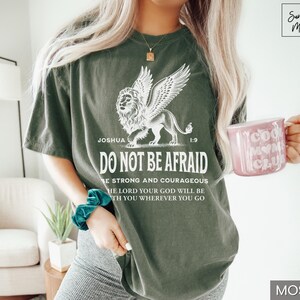 May include: Moss green t-shirt with a winged lion graphic and the text "DO NOT BE AFRAID". The shirt also features the text "Joshua 1:9" and other inspirational text. A pink mug with "Cool Mom Club" is held in the hand.