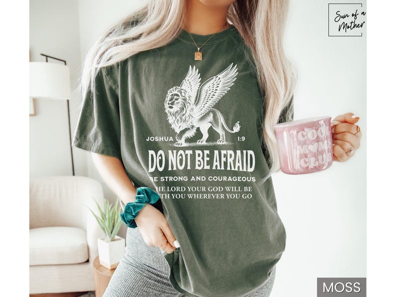 May include: Moss green t-shirt with a winged lion graphic and the text "DO NOT BE AFRAID". The shirt also features the text "Joshua 1:9" and other inspirational text. A pink mug with "Cool Mom Club" is held in the hand.