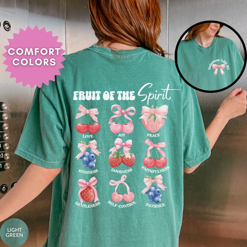 May include: A light green t-shirt with the text "FRUIT OF THE Spirit" in a retro font. The shirt features a graphic of nine different fruits with pink bows, each representing a different fruit of the spirit: love, joy, peace, kindness, goodness, faithfulness, gentleness, self-control, and patience.