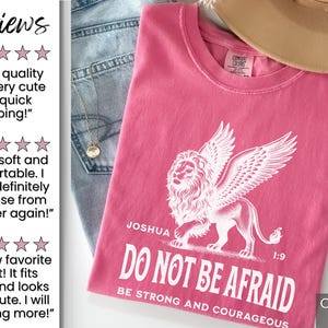 May include: A pink t-shirt with a white winged lion graphic and the text "DO NOT BE AFRAID". The shirt also has the text "Joshua 1:9" and "BE STRONG AND COURAGEOUS". The shirt is folded on top of blue jeans.