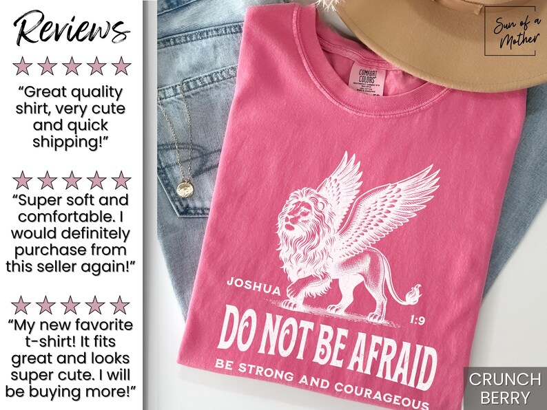 May include: A pink t-shirt with a white winged lion graphic and the text "DO NOT BE AFRAID". The shirt also has the text "Joshua 1:9" and "BE STRONG AND COURAGEOUS". The shirt is folded on top of blue jeans.