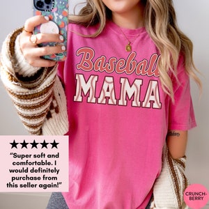 May include: Pink t-shirt with the text "Baseball Mama" in red and white lettering with a baseball stitch design. The text is in a vintage style font.