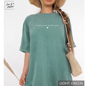 Puede incluir: Camiseta verde claro con el texto "reading is my favorite sport" y un peque&ntilde;o coraz&oacute;n. La camiseta es de color liso y tiene mangas cortas. El logotipo de la marca "Sun of a Mother" est&aacute; en la esquina superior izquierda.