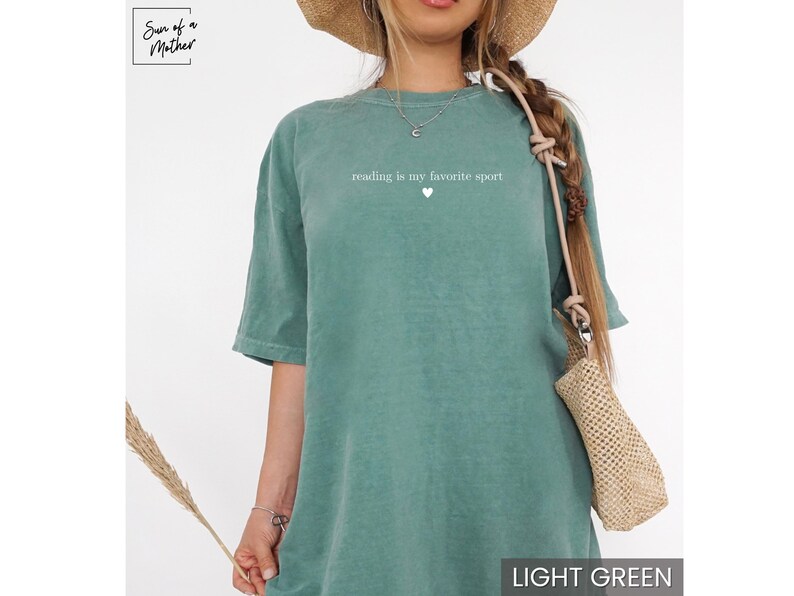 Puede incluir: Camiseta verde claro con el texto "reading is my favorite sport" y un peque&ntilde;o coraz&oacute;n. La camiseta es de color liso y tiene mangas cortas. El logotipo de la marca "Sun of a Mother" est&aacute; en la esquina superior izquierda.
