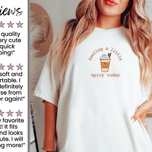 May include: White t-shirt with a fall-themed graphic. The design features a coffee cup with whipped cream, a cinnamon stick, and a pumpkin, with the text "feeling a little spicy today".
