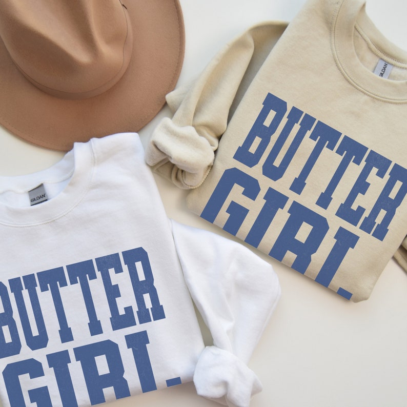 May include: Two sweatshirts with the text "Butter Girl" printed in blue on the front. One sweatshirt is white and the other is beige.