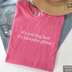 May include: A pink t-shirt with the text "it's not dog hair it's labrador glitter" in white. The shirt is folded and rests on a pair of blue jeans. A tan hat is on top of the shirt.
