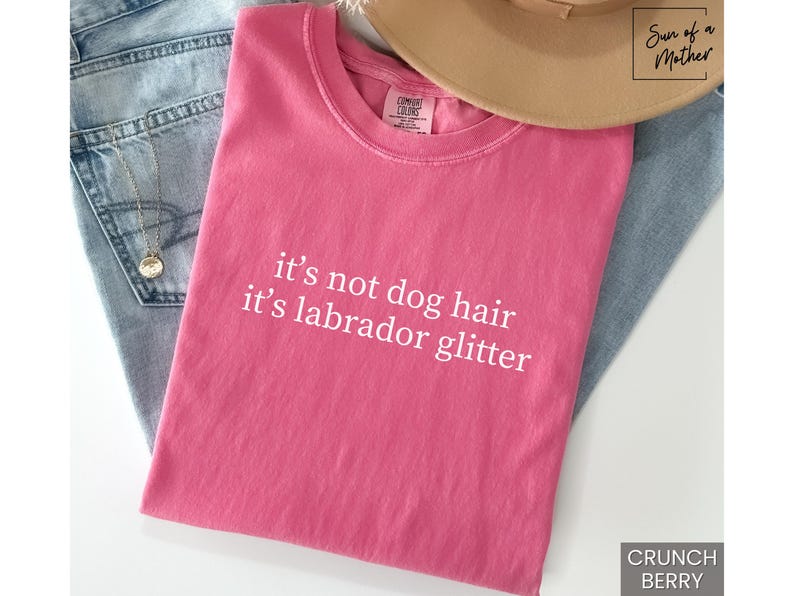 May include: A pink t-shirt with the text "it's not dog hair it's labrador glitter" in white. The shirt is folded and rests on a pair of blue jeans. A tan hat is on top of the shirt.