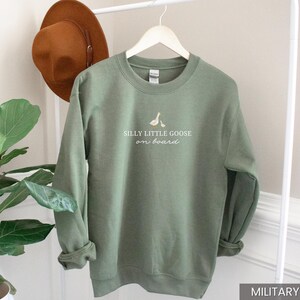 Peut inclure: Sweat-shirt vert militaire avec l'inscription blanche "Silly Little Goose on Board". Le sweat-shirt a un col rond et des manches longues. Un chapeau marron est suspendu &agrave; un portant blanc.