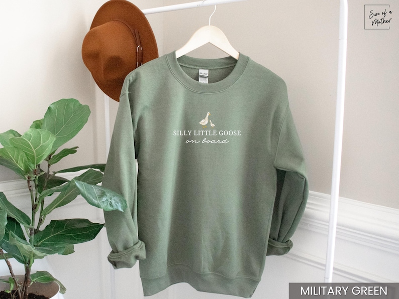 Peut inclure: Sweat-shirt vert militaire avec l'inscription blanche "Silly Little Goose on Board". Le sweat-shirt a un col rond et des manches longues. Un chapeau marron est suspendu &agrave; un portant blanc.
