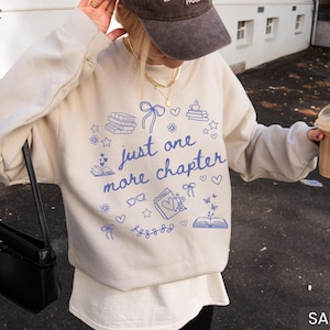 May include: Sand-colored sweatshirt with the text "just one more chapter" in blue, along with book and heart illustrations. The sweatshirt is a casual style, perfect for book lovers.