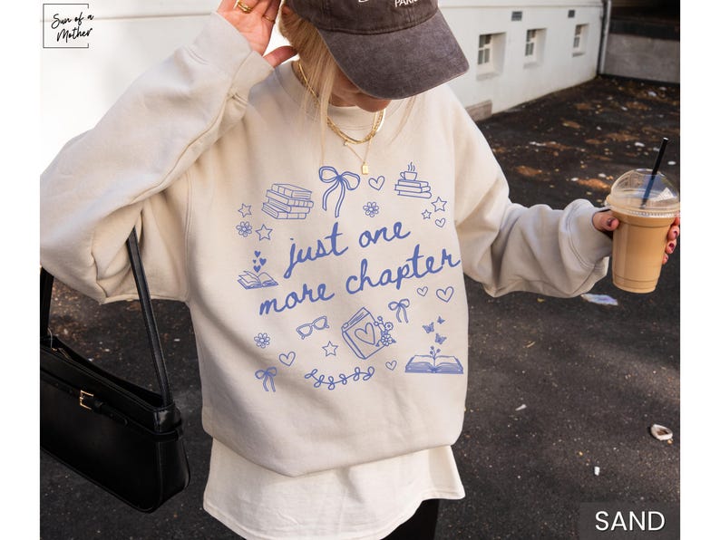 May include: Sand-colored sweatshirt with the text "just one more chapter" in blue, along with book and heart illustrations. The sweatshirt is a casual style, perfect for book lovers.