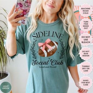以下が含まれることがあります： ターコイズブルーのTシャツに、フットボールのグラフィックと「Sideline Social Club Loud and Proud」の文字がプリントされています。