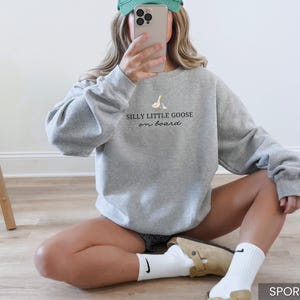 Peut inclure: Sweat-shirt gris sport avec l'inscription "Silly Little Goose on Board" et un petit dessin d'oie. Le sweat-shirt a des manches longues et un col rond. Le mod&egrave;le porte une casquette verte et des chaussettes blanches.