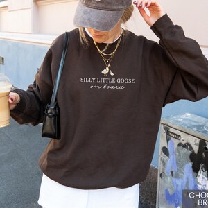 Peut inclure: Sweat-shirt marron chocolat avec l'inscription &laquo; Silly Little Goose on Board &raquo; et un dessin d'oie. Le sweat-shirt est de couleur unie et &agrave; manches longues. L'image montre &eacute;galement une personne portant une casquette et tenant une boisson au caf&eacute;.