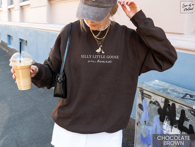 Peut inclure: Sweat-shirt marron chocolat avec l'inscription &laquo; Silly Little Goose on Board &raquo; et un dessin d'oie. Le sweat-shirt est de couleur unie et &agrave; manches longues. L'image montre &eacute;galement une personne portant une casquette et tenant une boisson au caf&eacute;.