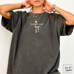 May include: Dark gray oversized t-shirt with the words "football mom" and a white bow graphic. The shirt has a vintage, worn look. The brand name "Sun of a Mother" is in the bottom right corner.