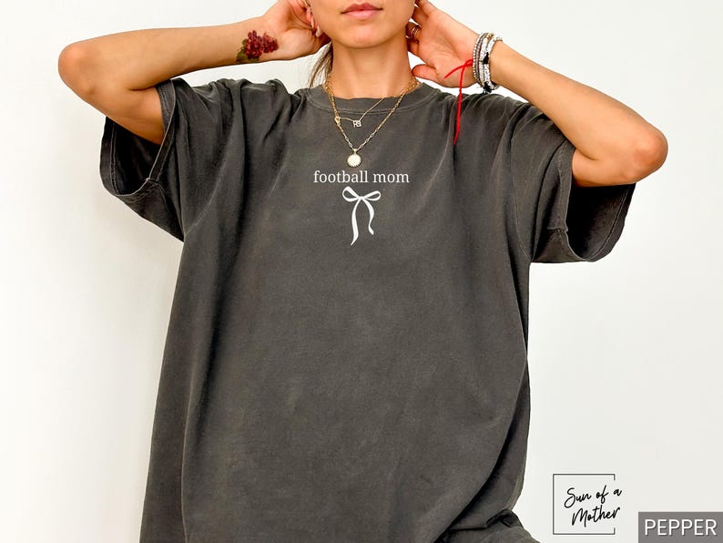 May include: Dark gray oversized t-shirt with the words "football mom" and a white bow graphic. The shirt has a vintage, worn look. The brand name "Sun of a Mother" is in the bottom right corner.