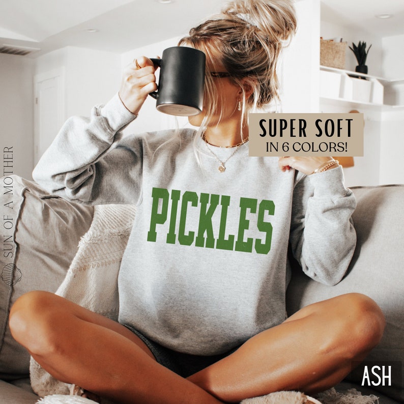 Op de afbeelding: Een grijs sweatshirt met het woord "PICKLES" bedrukt in groene letters. De tekst "SUPER SOFT IN 6 COLORS!" is in bruine letters gedrukt aan de rechterkant van het sweatshirt. Het sweatshirt wordt gedragen door een persoon die op een bank zit.
