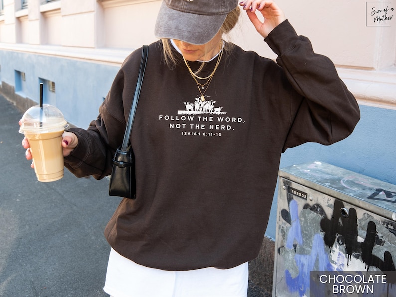 May include: Chocolate brown sweatshirt with the text "FOLLOW THE WORD, NOT THE HERD" and the Bible verse Isaiah 8:11-13 in white. The sweatshirt has a crew neck and long sleeves. The image also shows a person wearing a gray cap and holding a coffee drink.
