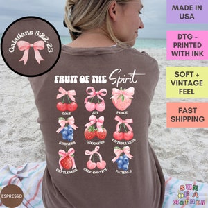 May include: A brown t-shirt with the text "Fruit of the Spirit" and a graphic of nine fruits with pink bows. The fruits are cherries, strawberries, and blueberries. The text "Galatians 5:22-23" is on a pink bow in the top left corner.
