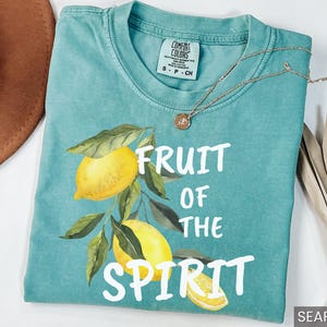 Op de afbeelding: Een tealblauw t-shirt met een witte afbeelding van citroenen en bladeren. De tekst "Fruit of the Spirit" is in wit op het shirt gedrukt.