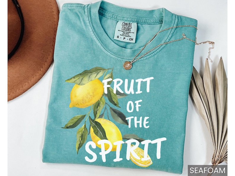Op de afbeelding: Een tealblauw t-shirt met een witte afbeelding van citroenen en bladeren. De tekst "Fruit of the Spirit" is in wit op het shirt gedrukt.