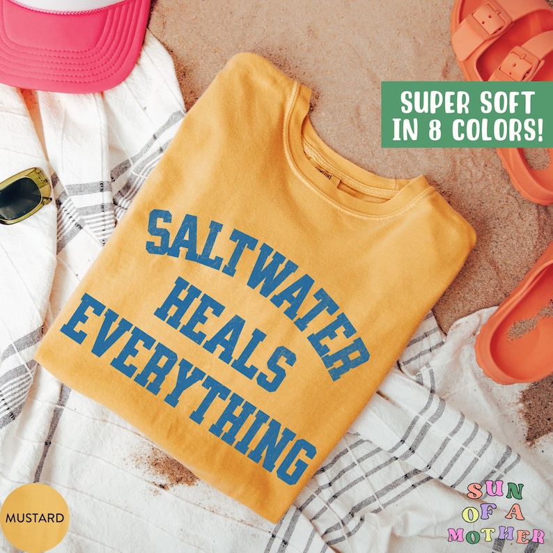 May include: A mustard yellow t-shirt with the text "Saltwater Heals Everything" in blue lettering. The t-shirt is folded and lying on a white and gray striped towel on a sandy surface. The text "Super Soft in 8 Colors!" is in green text on a white background. The text "Sun of a Mother" is in rainbow lettering on a white background.