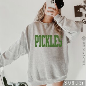 Op de afbeelding: Een grijs sweatshirt met het woord "PICKLES" in groene letters.