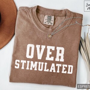 May include: A brown t-shirt with the text "OVER STIMULATED" in white lettering. The shirt is made of a soft, comfortable material and has a relaxed fit.