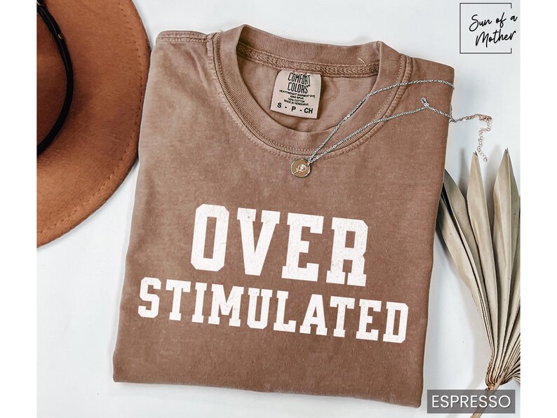 May include: A brown t-shirt with the text "OVER STIMULATED" in white lettering. The shirt is made of a soft, comfortable material and has a relaxed fit.