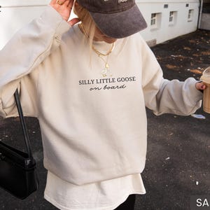 Peut inclure: Sweat-shirt couleur sable avec l'inscription "SILLY LITTLE GOOSE on board" en &eacute;criture noire. Le sweat-shirt est associ&eacute; &agrave; un sous-v&ecirc;tement blanc et &agrave; un sac &agrave; main noir. La personne porte une casquette marron et tient une boisson au caf&eacute;.
