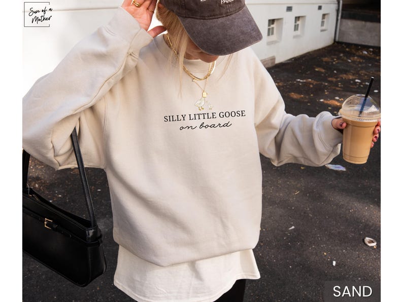 Peut inclure: Sweat-shirt couleur sable avec l'inscription "SILLY LITTLE GOOSE on board" en &eacute;criture noire. Le sweat-shirt est associ&eacute; &agrave; un sous-v&ecirc;tement blanc et &agrave; un sac &agrave; main noir. La personne porte une casquette marron et tient une boisson au caf&eacute;.