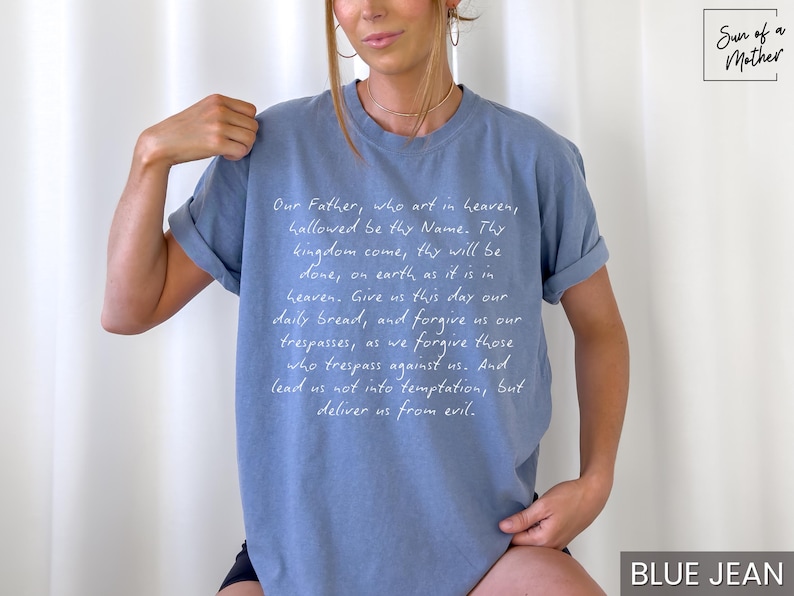 May include: A light blue t-shirt with white text that reads "Our Father, who art in heaven, hallowed be thy Name. Thy kingdom come, thy will be done, on earth as it is in heaven. Give us this day our daily bread, and forgive us our trespasses, as we forgive those who trespass against us. And lead us not into temptation, but deliver us from evil." The text is written in a cursive font. The shirt has a crew neck and short sleeves.