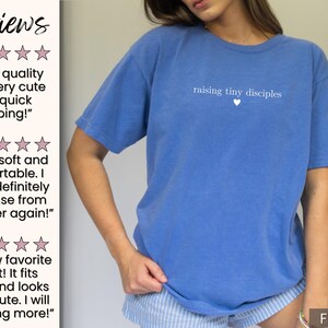 Op de afbeelding: Een blauw t-shirt met de tekst "raising tiny disciples" en een klein hartje. Het shirt is effen van kleur en heeft korte mouwen. Het shirt heeft de kleur Flo Blu.