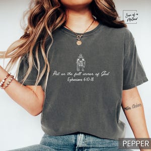 Puede incluir: Una camiseta gris oscuro con un gr&aacute;fico blanco de una persona con armadura y el texto "Ponte la armadura completa de Dios Efesios 6:10-18".