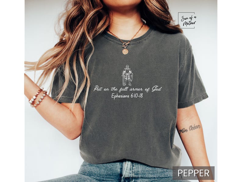 Puede incluir: Una camiseta gris oscuro con un gr&aacute;fico blanco de una persona con armadura y el texto "Ponte la armadura completa de Dios Efesios 6:10-18".
