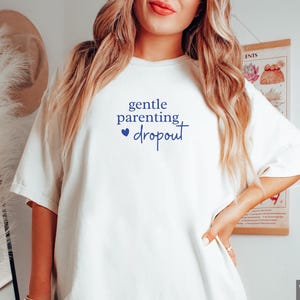 Puede incluir: Camiseta blanca con el texto "gentle parenting dropout" en azul marino. El texto incluye un peque&ntilde;o coraz&oacute;n azul. La camiseta tiene un corte holgado y mangas cortas. Prenda de vestir informal.