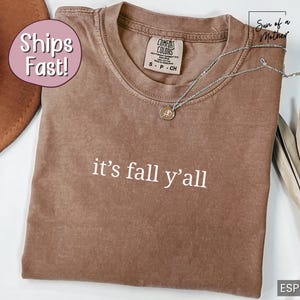May include: Espresso-colored Comfort Colors t-shirt with the phrase "it's fall y'all" in white lettering. A silver necklace with a pendant rests on the shirt. The shirt is folded, and a brown hat is partially visible.