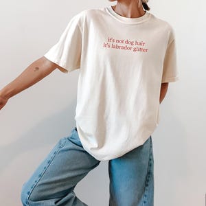 May include: Ivory-colored t-shirt with the text "it's not dog hair it's labrador glitter" in red. The shirt is paired with light blue jeans. The overall style is casual and comfortable.