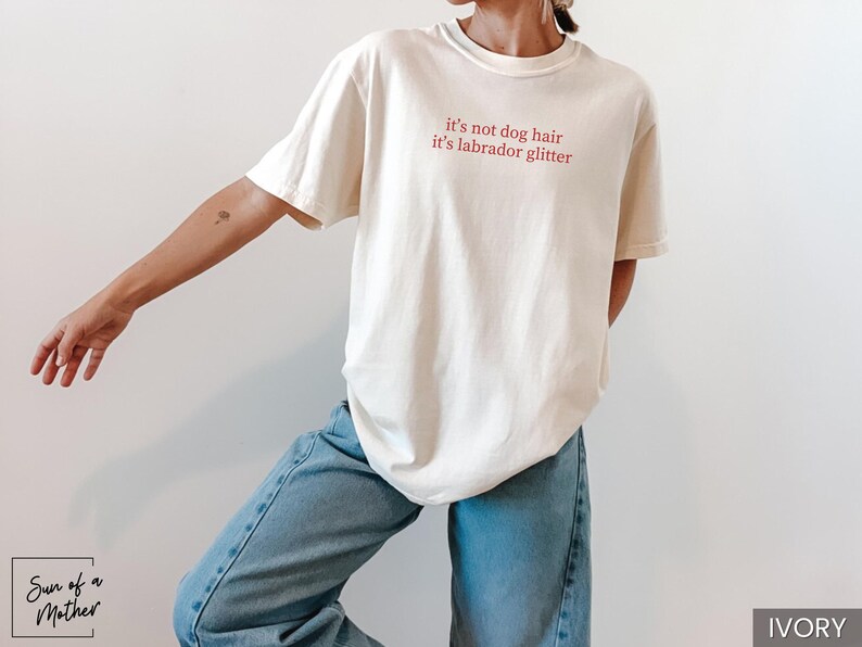 May include: Ivory-colored t-shirt with the text "it's not dog hair it's labrador glitter" in red. The shirt is paired with light blue jeans. The overall style is casual and comfortable.