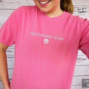 以下が含まれることがあります： ピンク色のTシャツ。白い文字で「dachshund mom」と足跡のマークがプリントされています。クルーネックで無地です。「Crunch-Berry」の文字が右下にあります。