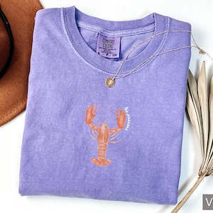 Może przedstawiać: Fioletowy t-shirt z grafiką homara i tekstem "be fearless".