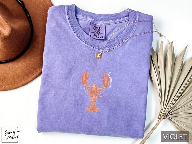 Może przedstawiać: Fioletowy t-shirt z grafiką homara i tekstem "be fearless".