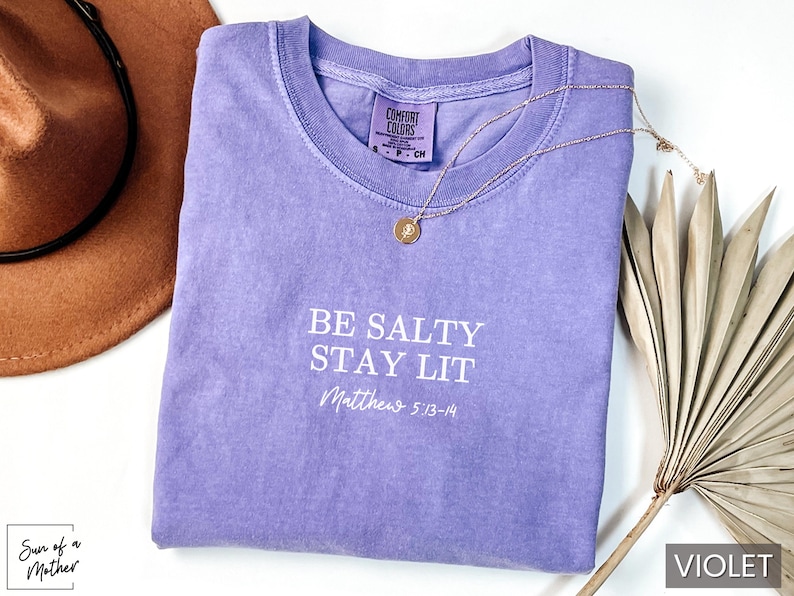 Puede incluir: Una camiseta morada con el texto "BE SALTY STAY LIT" y "Matthew 5:13-14" en letras blancas.