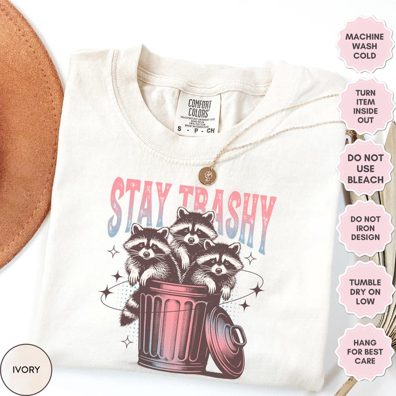 Op de afbeelding: Een beige T-shirt met de tekst "Stay Trashy" in roze en blauwe letters. De afbeelding toont drie wasberen in een prullenbak met sterren eromheen. Het shirt heeft wasinstructies aan de rechterkant.
