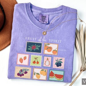 May include: Purple t-shirt with a graphic of fruit stamps. The stamps feature different fruits, including strawberries, cherries, grapes, and a pineapple. The text "FRUIT of the SPIRIT" is printed above the stamps.