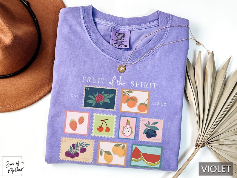 May include: Purple t-shirt with a graphic of fruit stamps. The stamps feature different fruits, including strawberries, cherries, grapes, and a pineapple. The text "FRUIT of the SPIRIT" is printed above the stamps.