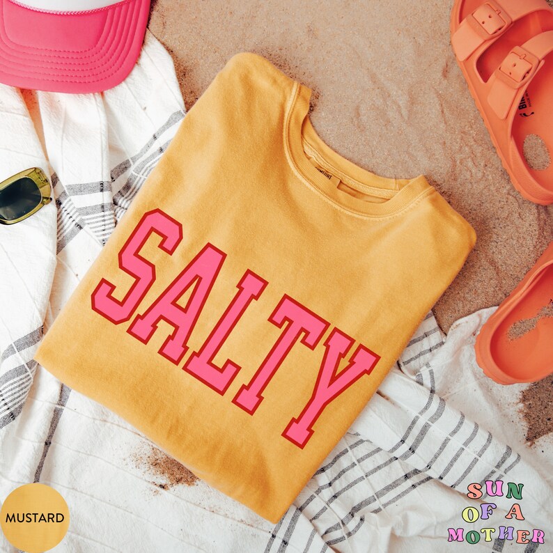 May include: A mustard yellow t-shirt with the word "SALTY" printed in pink letters. The shirt is folded and lying on a white and gray striped towel. The background is a sandy surface.